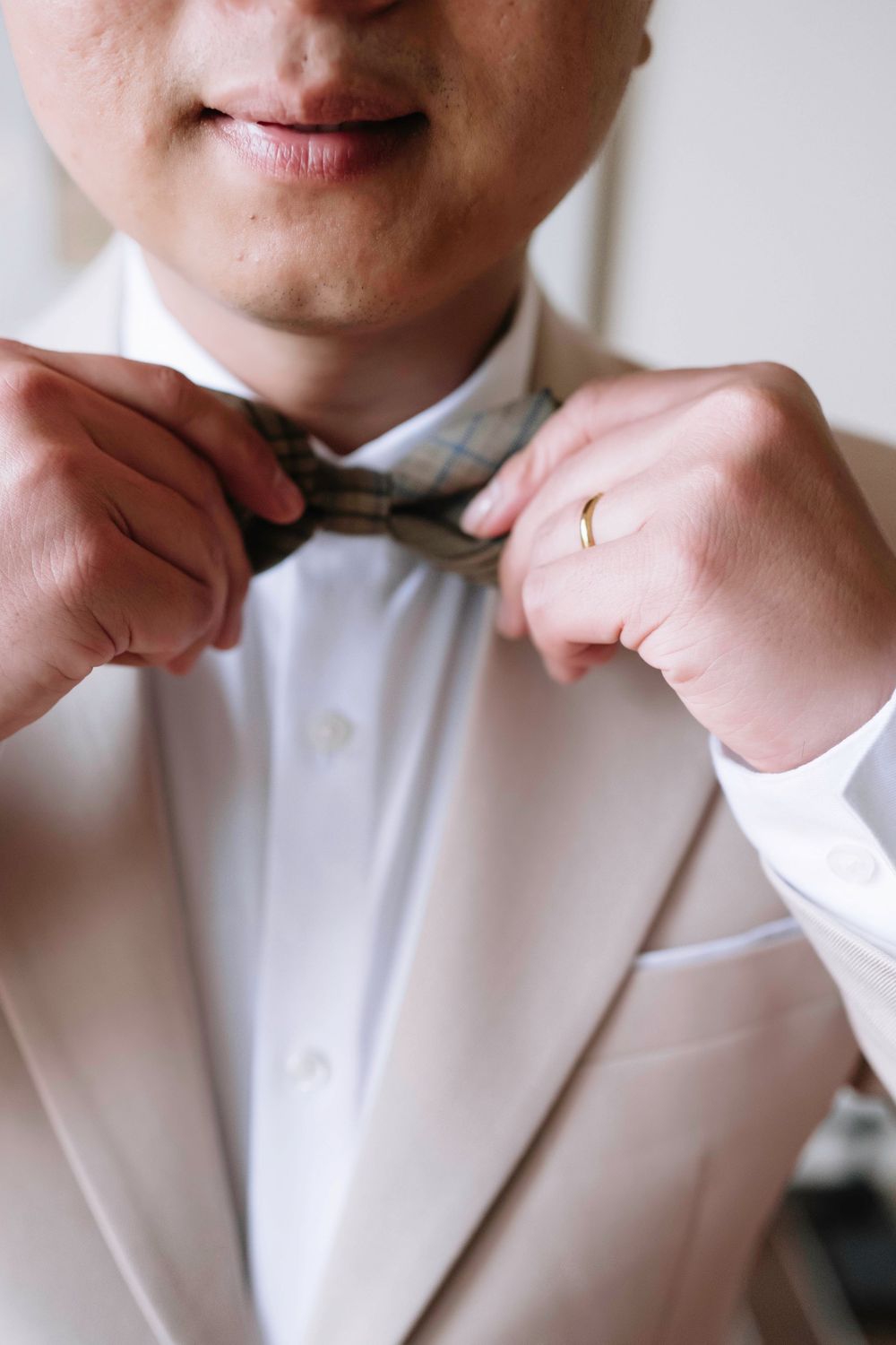 Groom in beige suit adjusting his bow tie with a gold wedding band on his finger.