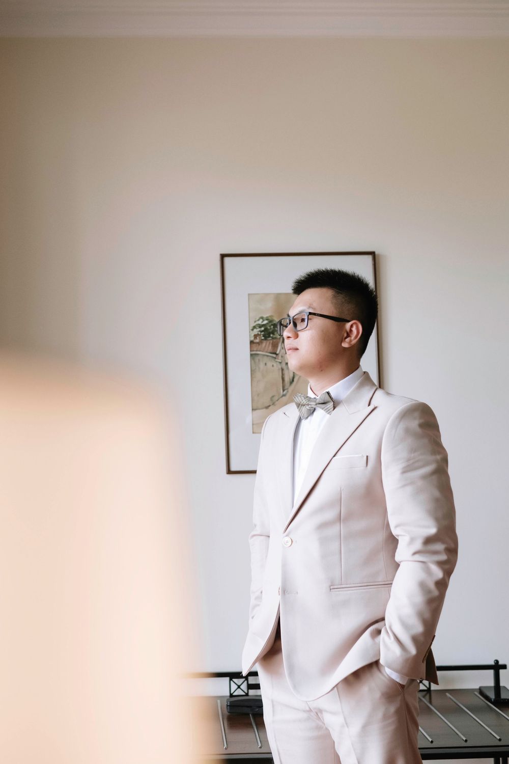 Groom in beige suit and bow tie standing thoughtfully in elegant room before wedding ceremony.