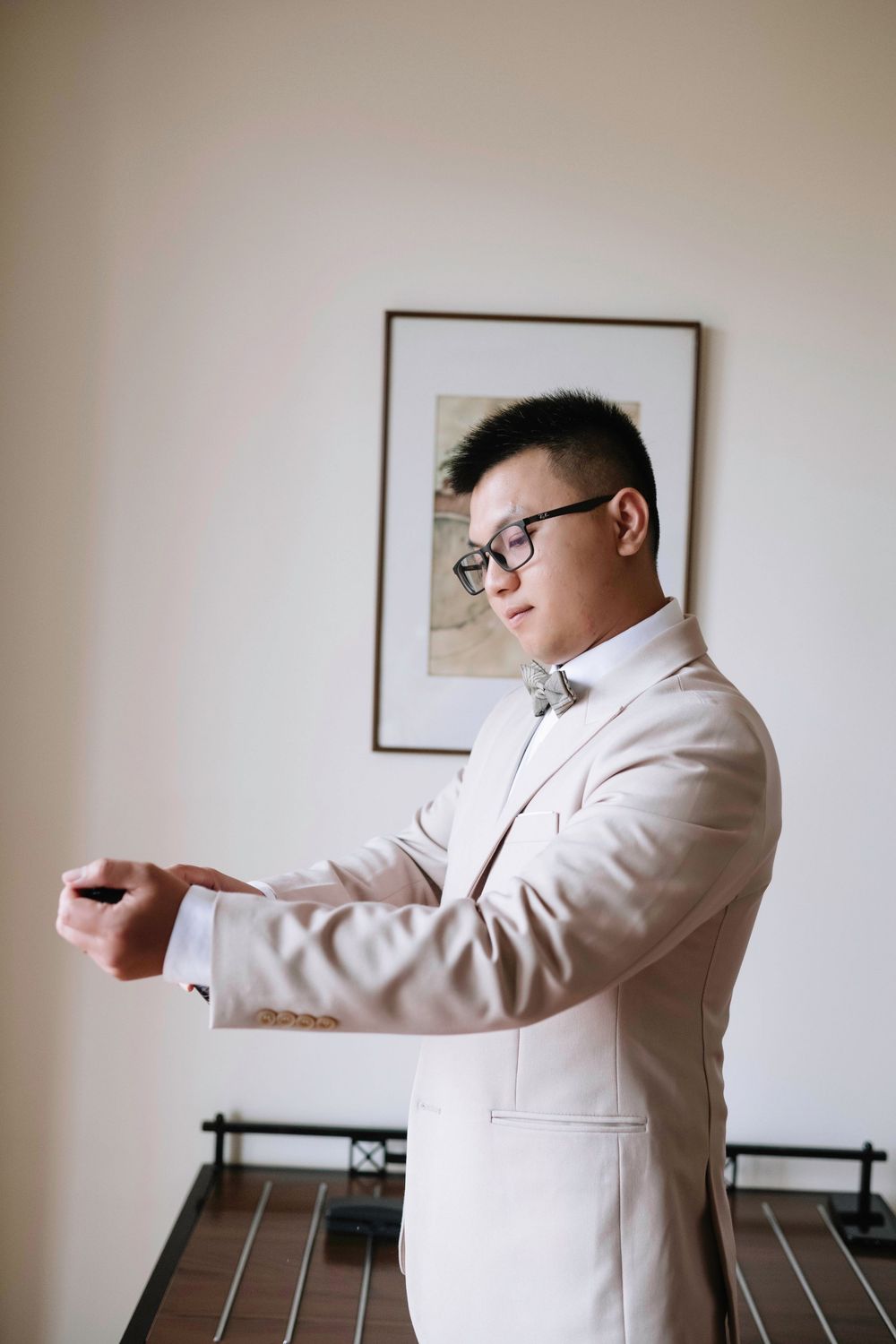 Groom in cream suit adjusting cufflinks while preparing for wedding day indoors.