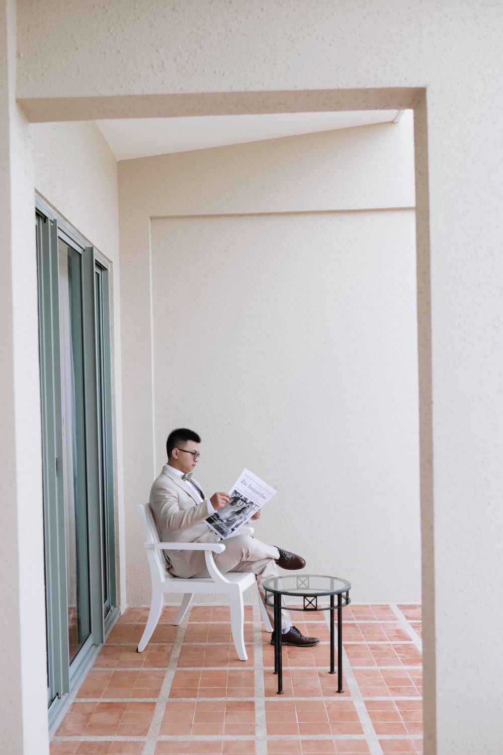 Groom in beige suit reading newspaper on hotel balcony before wedding ceremony.