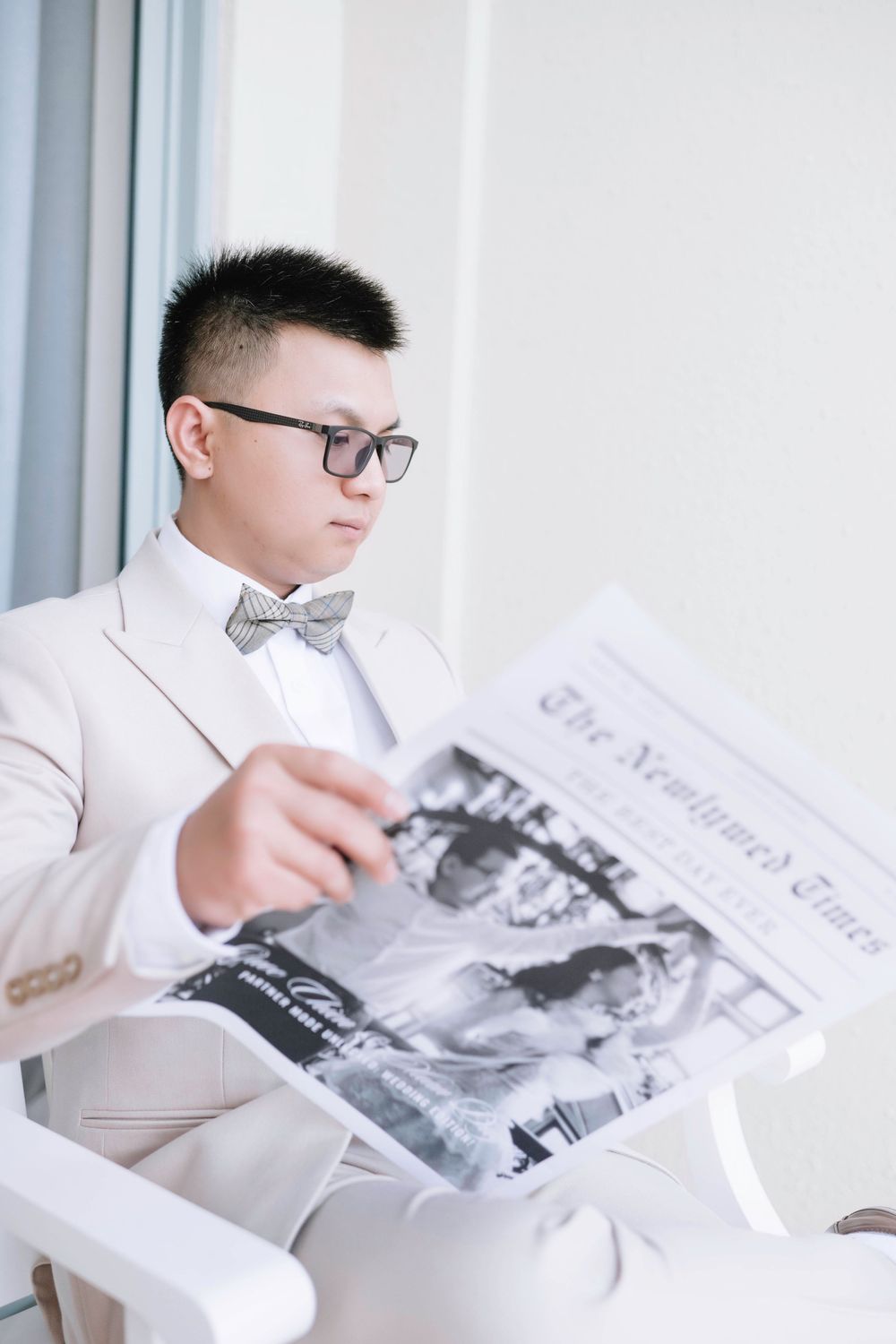 Groom in ivory suit reading The Newlywed Times newspaper while seated in white chair.