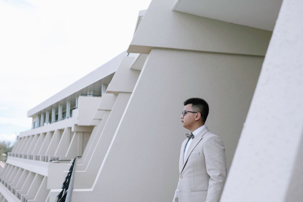 Young Asian man in light gray suit standing by modern white architectural building exterior.