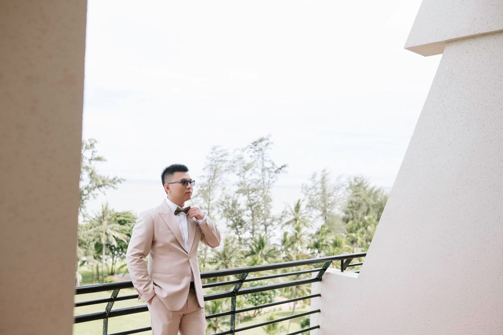 Groom in beige suit adjusting bow tie on outdoor balcony with tropical greenery backdrop.