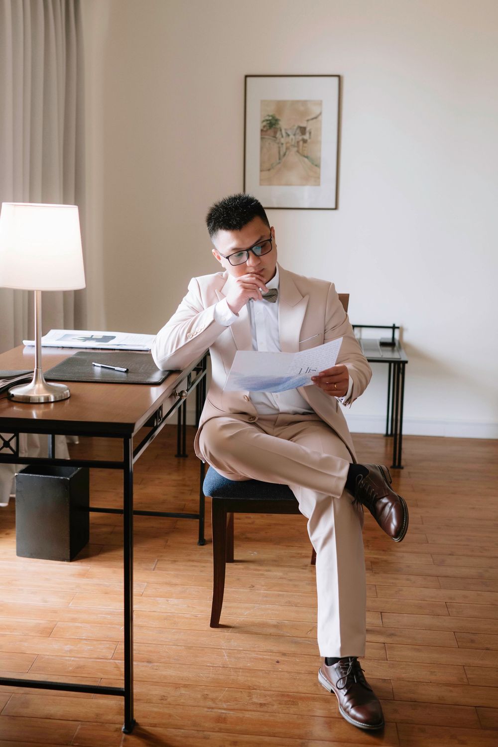 Groom in cream suit reviewing wedding vows while seated at desk in hotel room.
