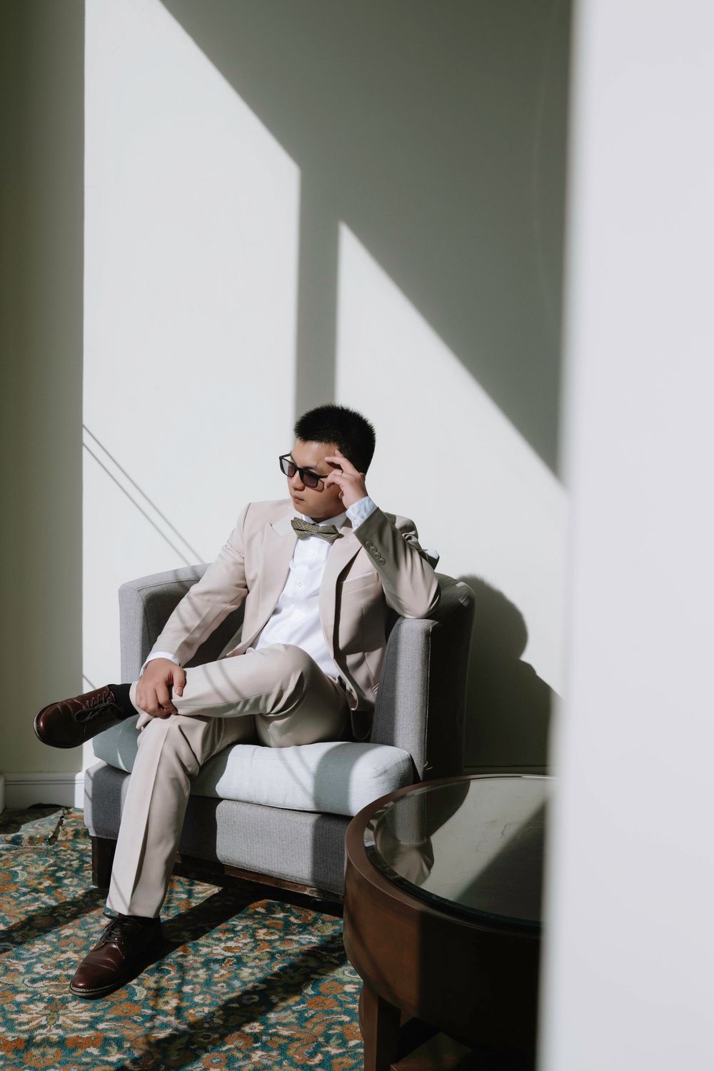 Groom in beige suit and sunglasses relaxing in armchair with dramatic sunlight shadows.