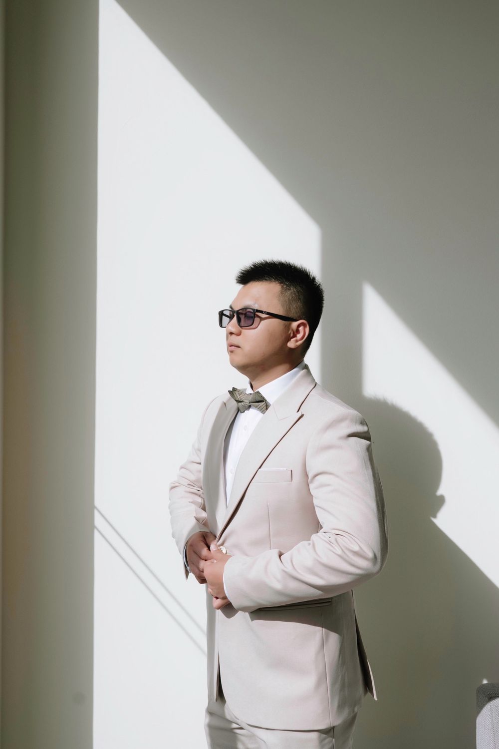 Groom in ivory suit and bow tie standing by sunlit white wall before ceremony.