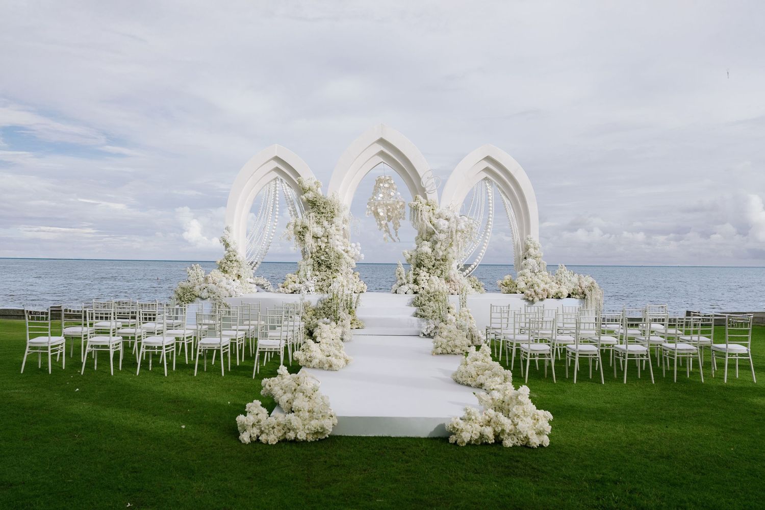 All-white outdoor wedding ceremony setup with floral arches and chairs facing the ocean.