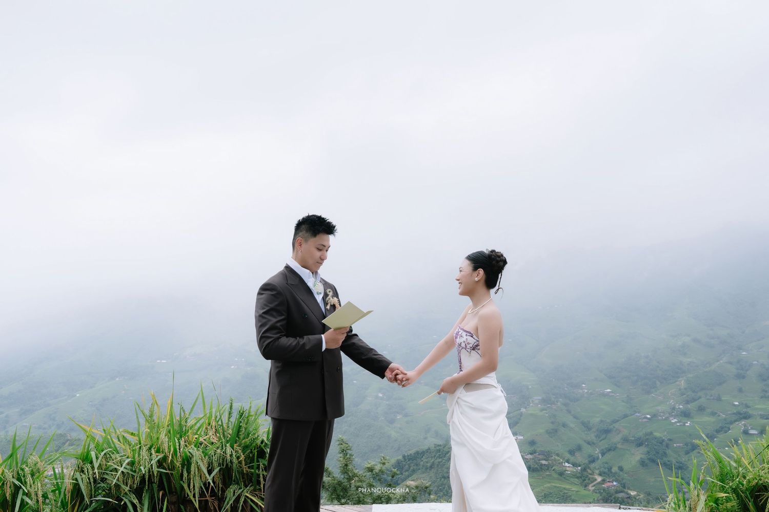 Groom reading vows to bride while holding hands on misty mountain overlook during outdoor ceremony.