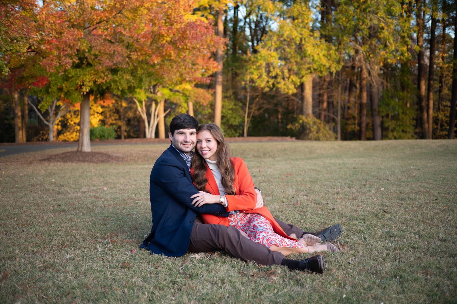 A couple sitting together on grass surrounded by vibrant fall foliage and trees displaying autumn colors.