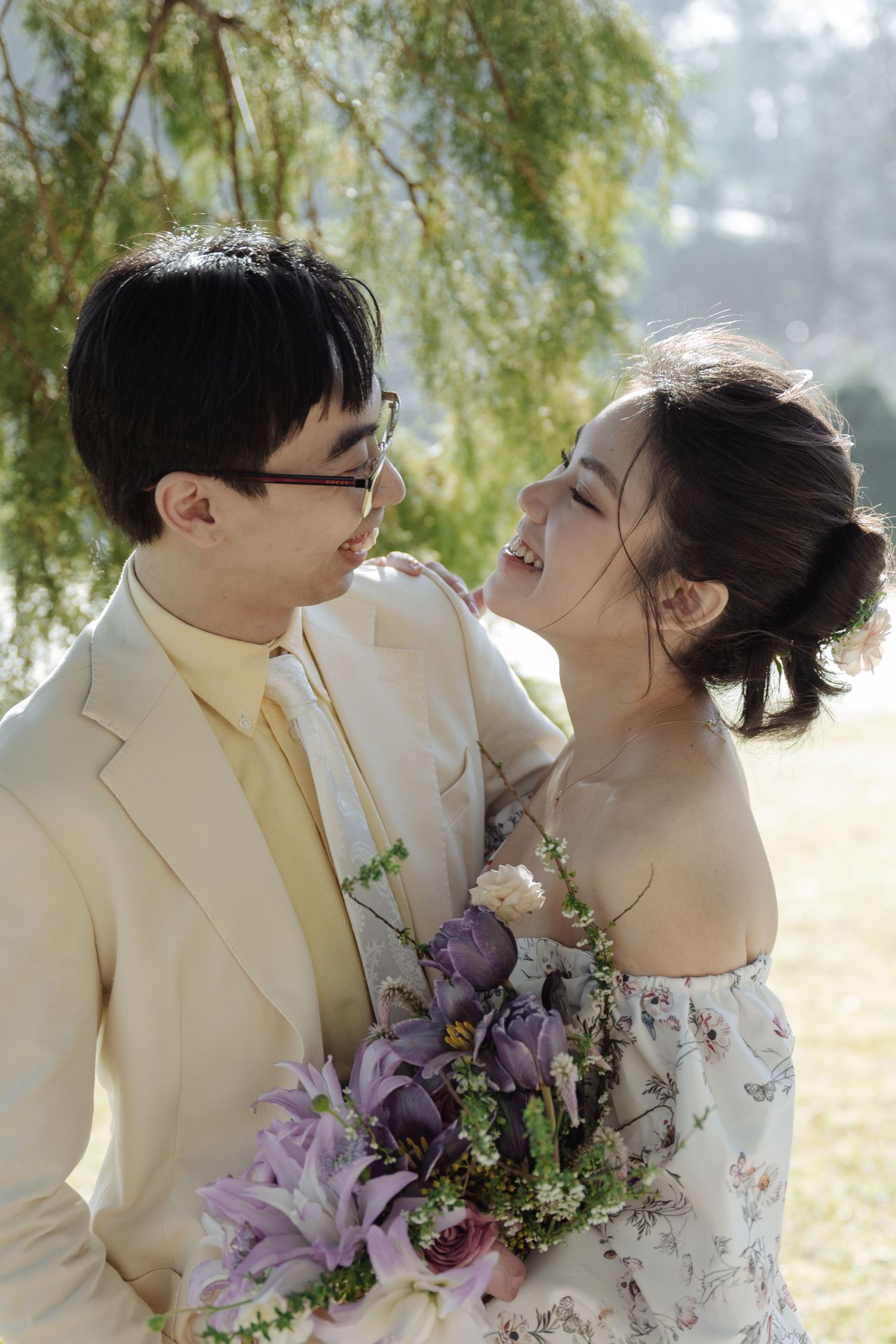 Joyful Asian couple in garden setting, groom in cream suit holding purple bouquet while bride laughs in floral dress.
