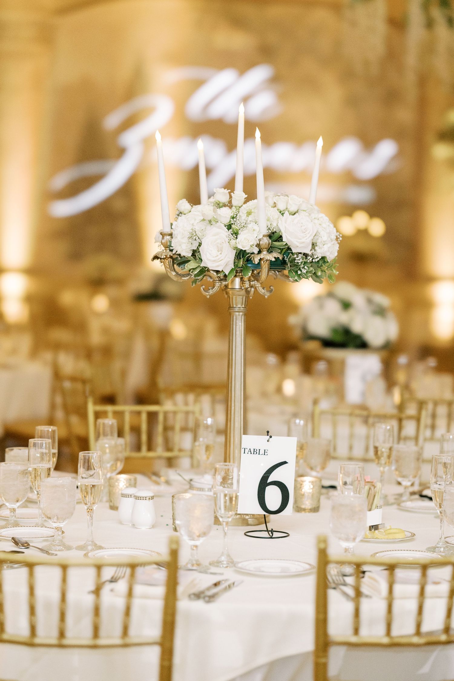 Elegant wedding reception tables with white floral centerpieces and gold chairs in a warmly lit ballroom setting.
