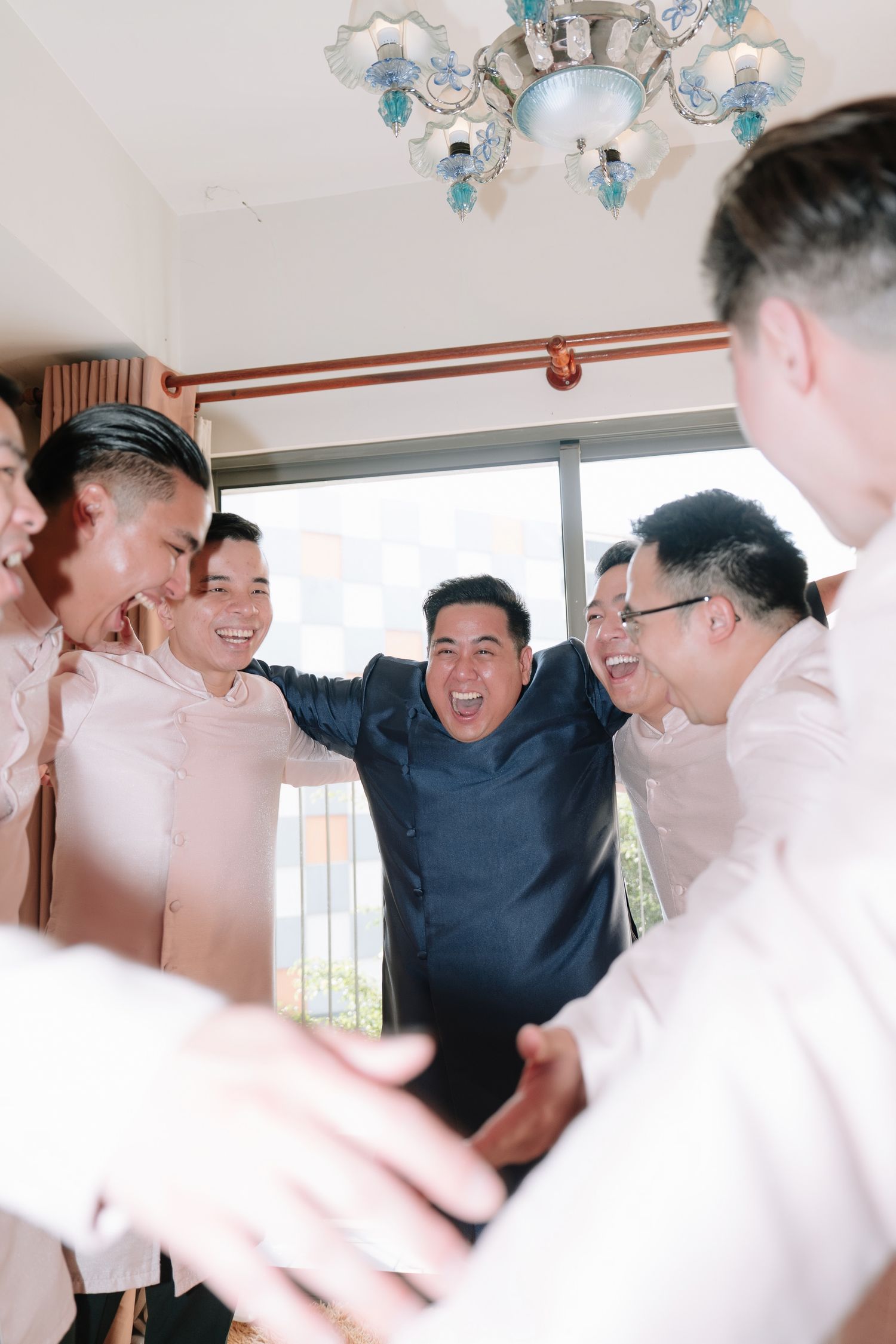 Joyful groom in navy ao dai laughing with groomsmen in pink traditional Vietnamese outfits indoors.