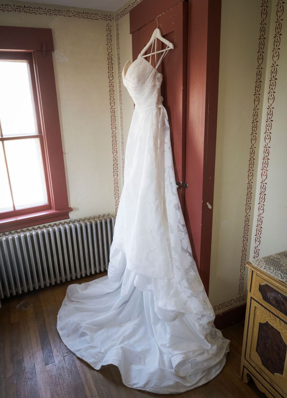 A white wedding dress hangs elegantly by a window in a vintage-style room.