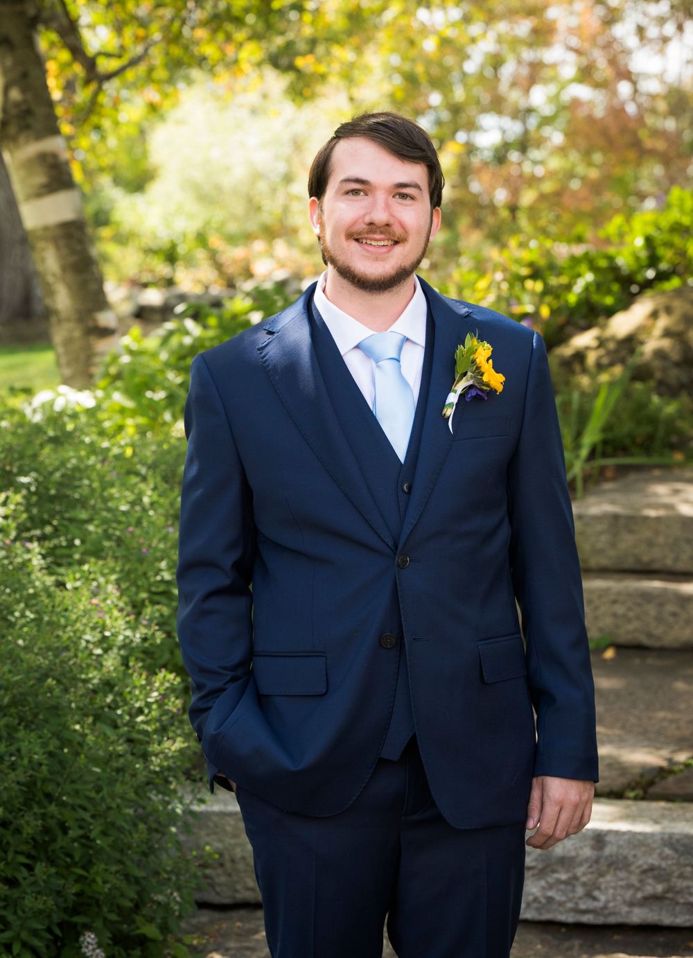 Wedding party member in a navy suit with sunflower boutonniere standing outdoors.