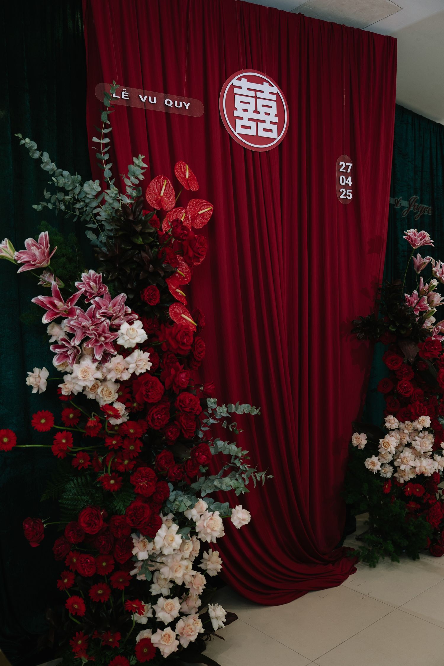 Red curtain wedding backdrop with double happiness symbol, floral arrangements, and date 27.04.25.