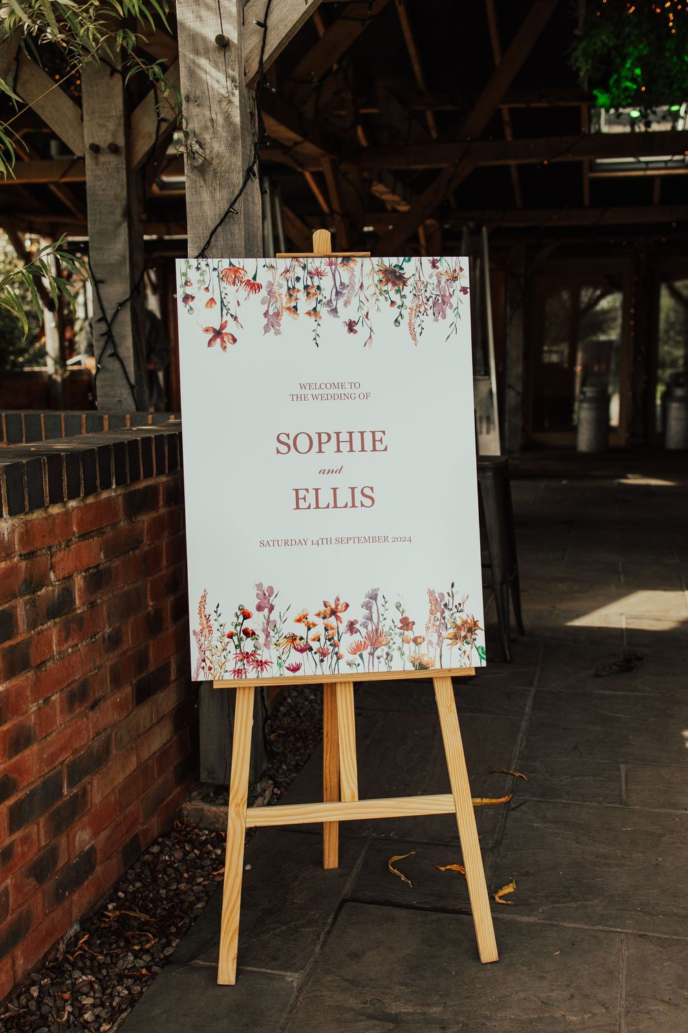 White wedding signs with floral designs displayed on wooden easels in a rustic brick venue.