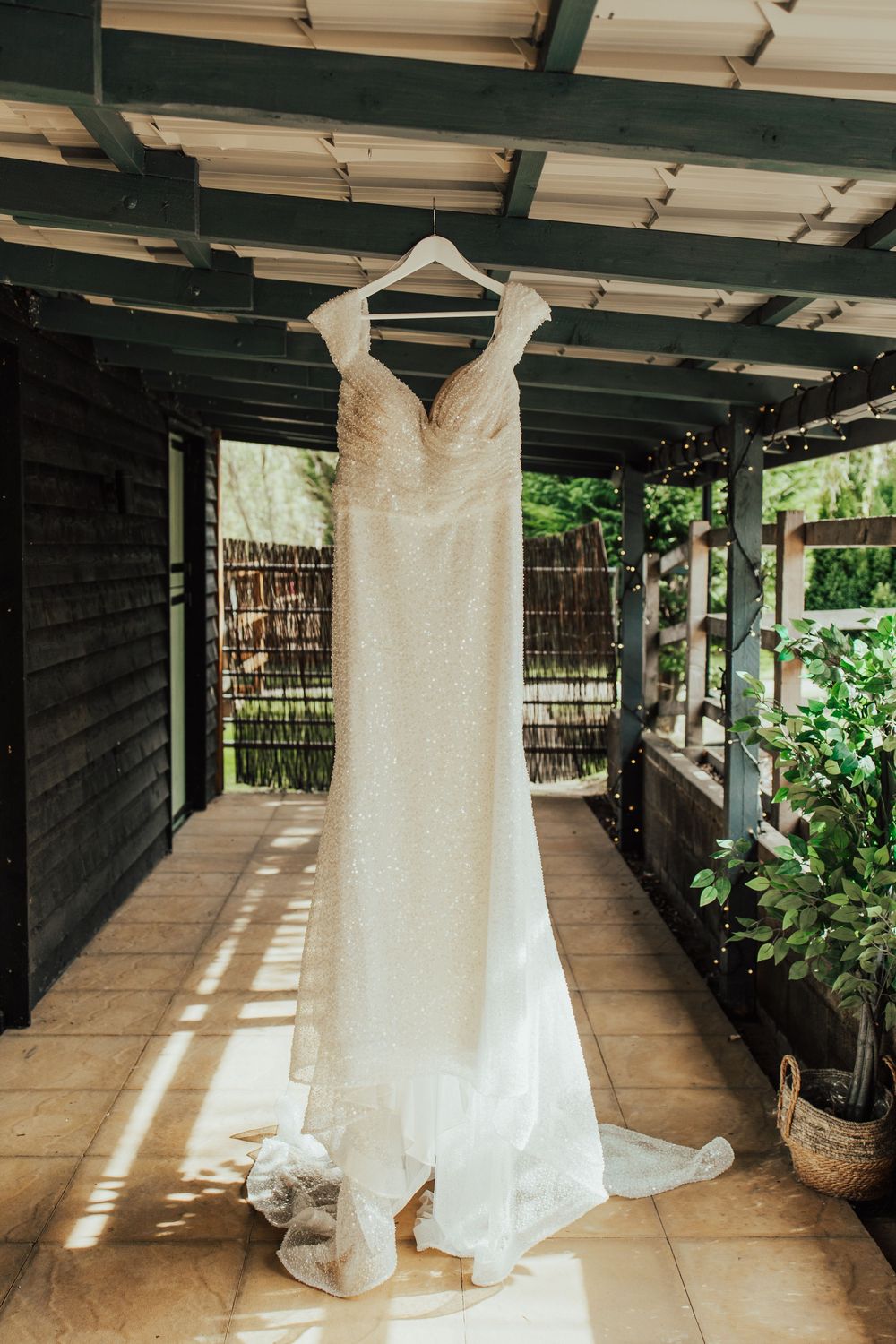 A white lace wedding dress hangs elegantly on a wooden porch with sunlight streaming through.