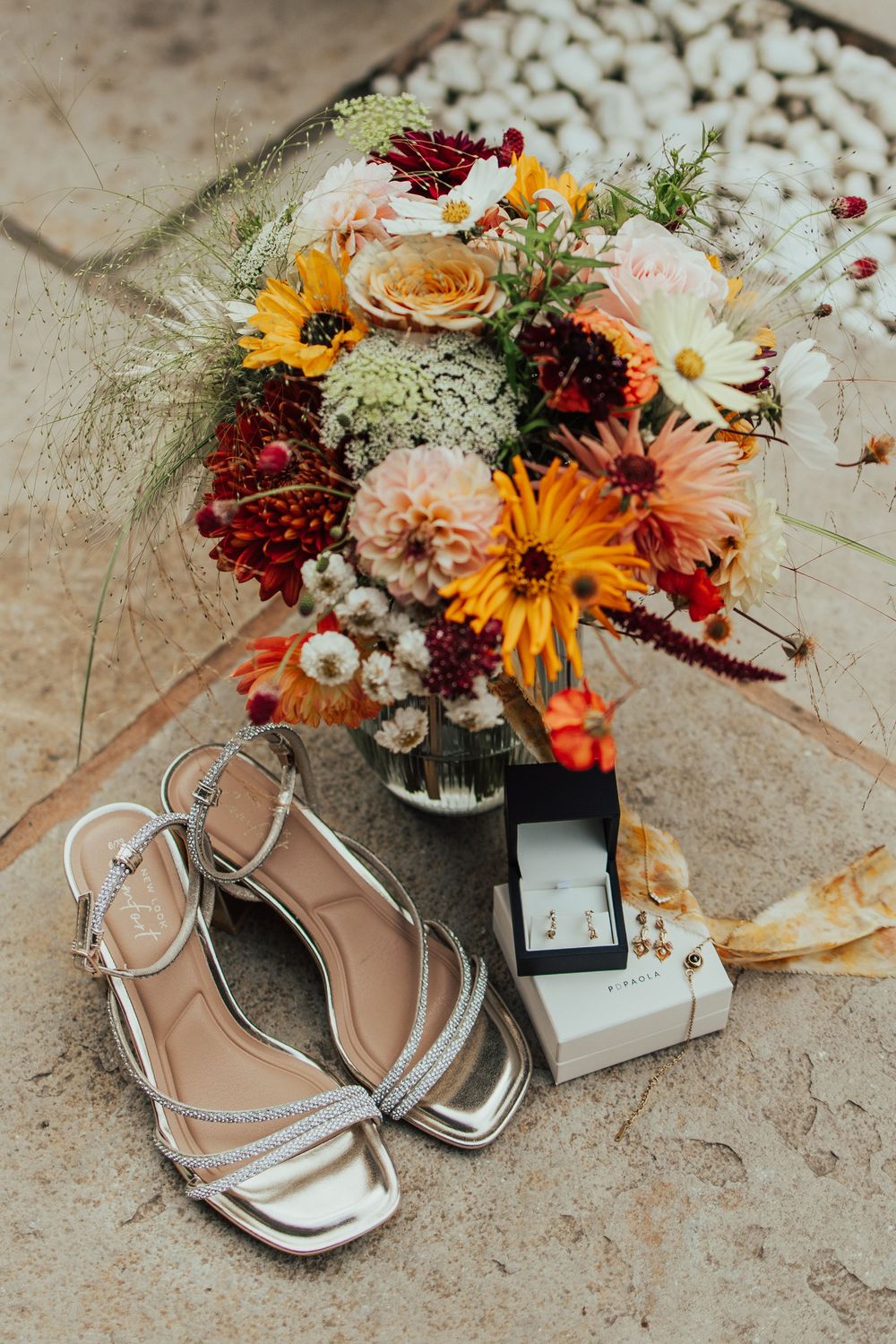 A colorful wedding bouquet with sunflowers and roses next to silver sandals and a ring box on a beige surface.