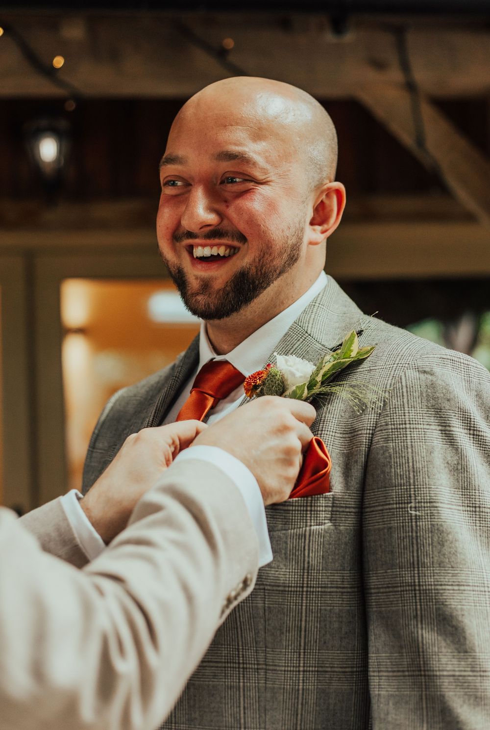 Groomsman in gray plaid suit and red tie getting boutonniere pinned to lapel during wedding preparations.