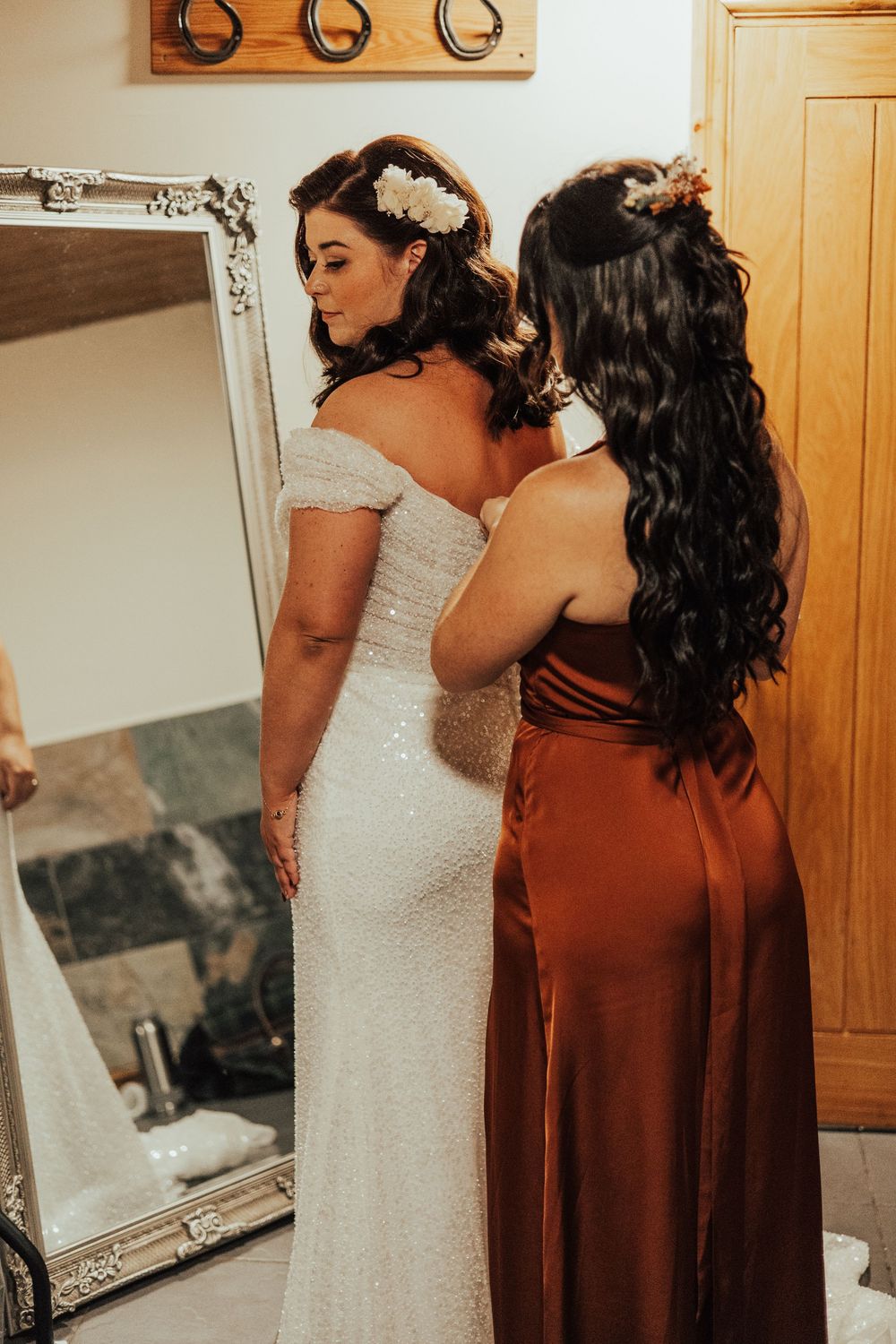 A bride in a white lace gown and bridesmaid in brown dress share a quiet moment before the wedding.