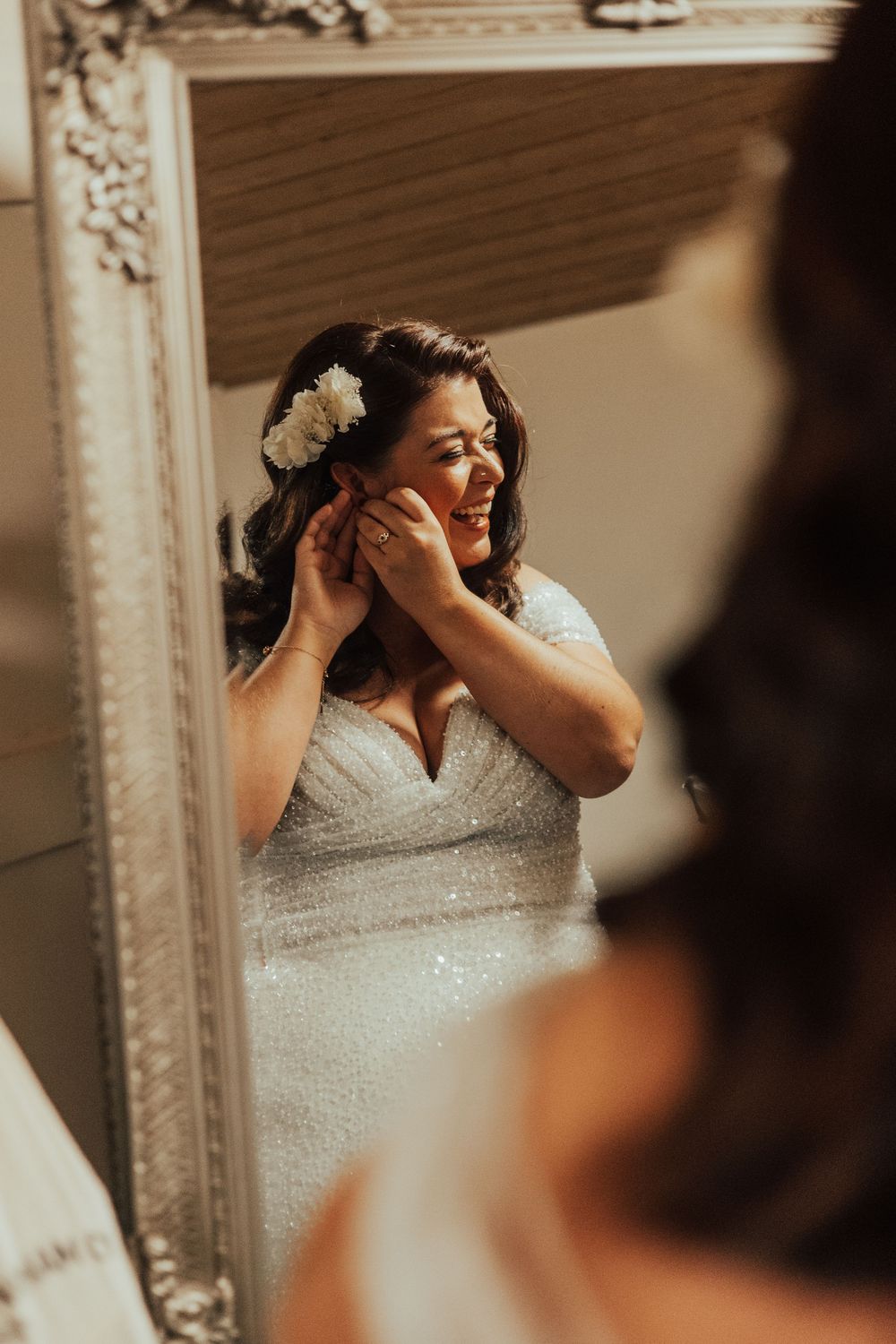Wedding preparation scene showing a bride in a beaded white dress adjusting her headpiece in a decorative mirror.