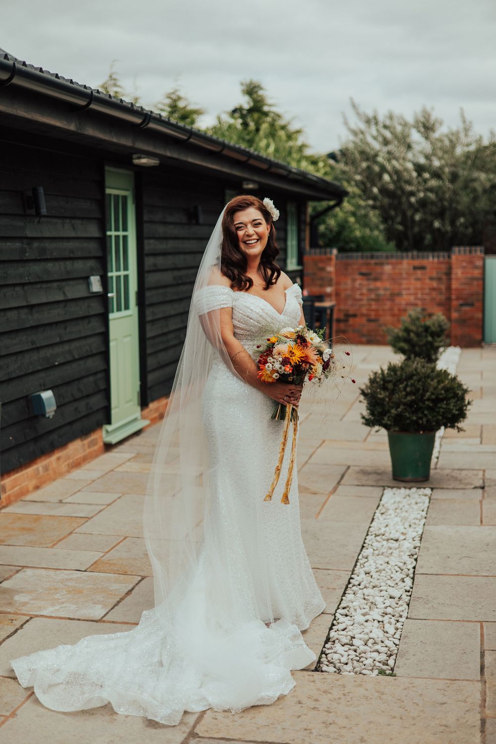 An elegant bride in a white wedding gown with a flowing veil twirls on a stone patio next to a brick and wood building.