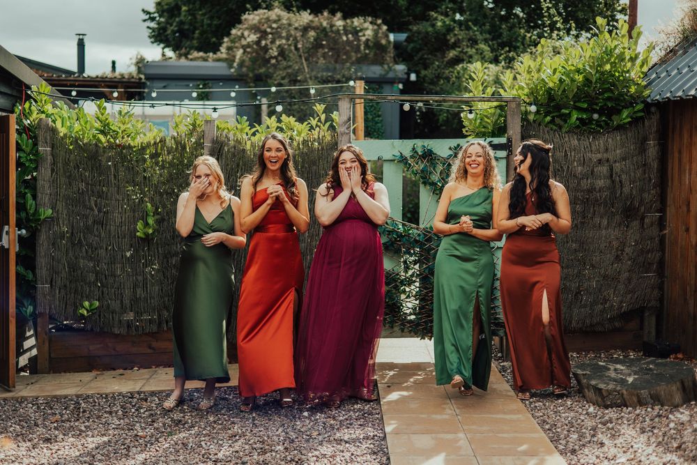 A group of bridesmaids in jewel-toned dresses pose together in an outdoor garden setting.