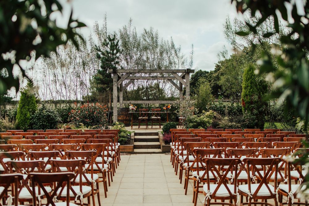 Brown wooden chairs arranged in rows outdoors for a wedding ceremony with greenery and trees in the background.