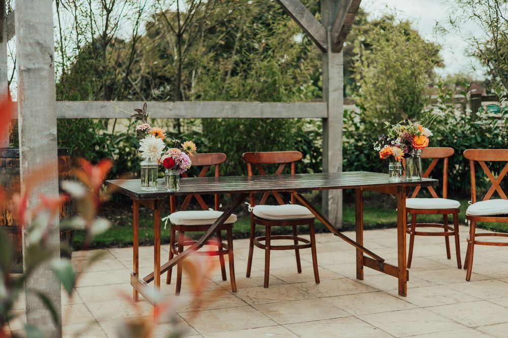 Empty wooden chairs and tables arranged on an outdoor patio with greenery and orange flowers in the background.