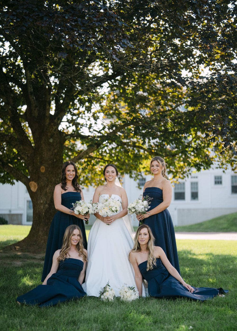 A wedding party poses on a grassy lawn under a large tree wearing navy blue dresses and a white gown.