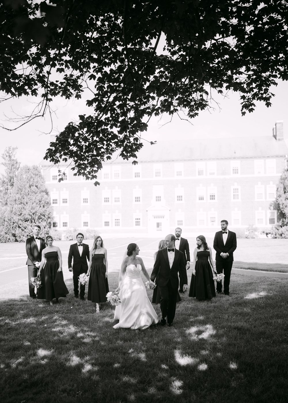 A wedding party stands in a row on a grassy field with a building in the background.