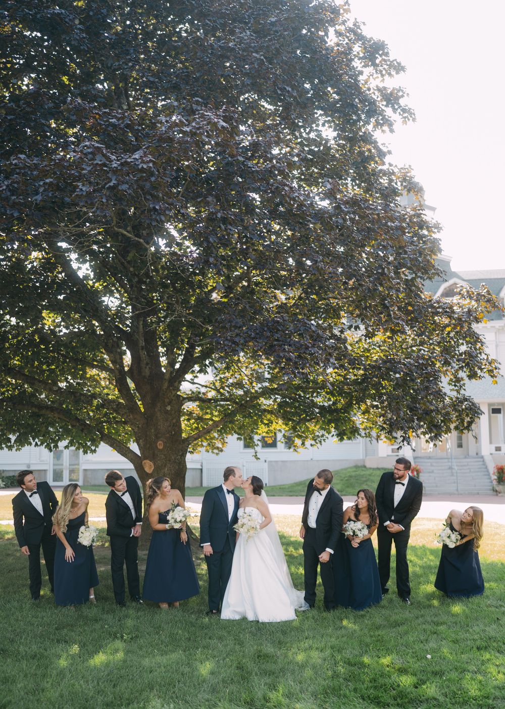 Wedding party lined up under large tree for outdoor group photo.