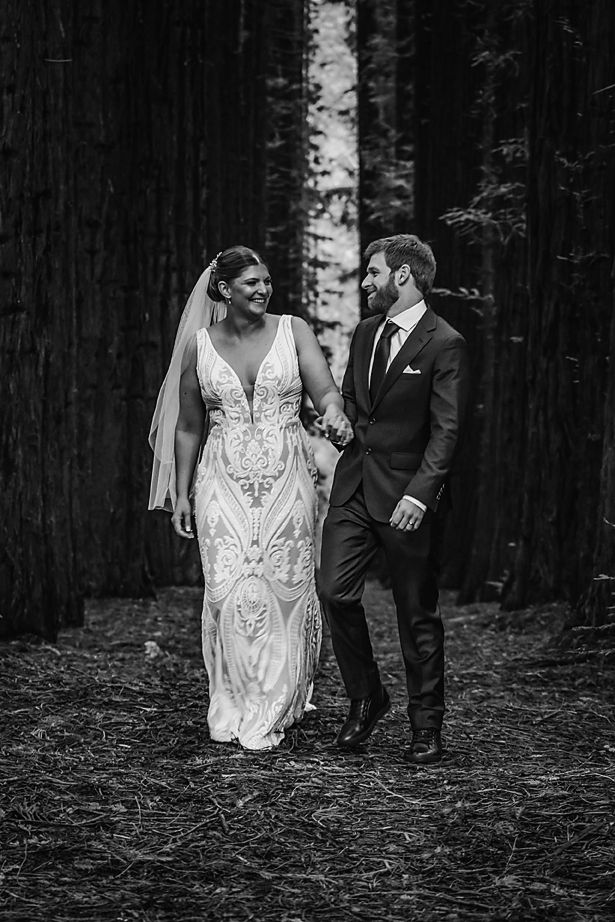 Black and white wedding photography of a couple in formal attire walking through a forest of birch trees.