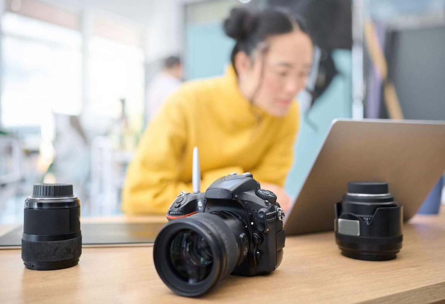 A series of camera lenses and equipment arranged in a row on a wooden desk surface with computers in the background.