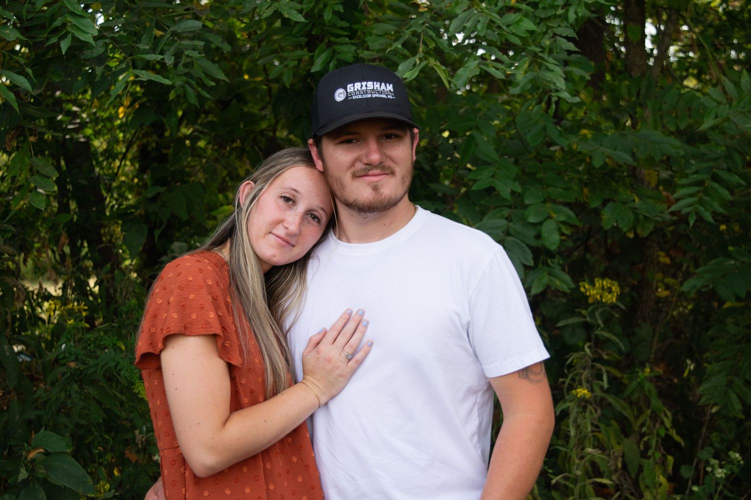 Couple standing in front of full trees hugging, woman has her head resting on man's shoulder
