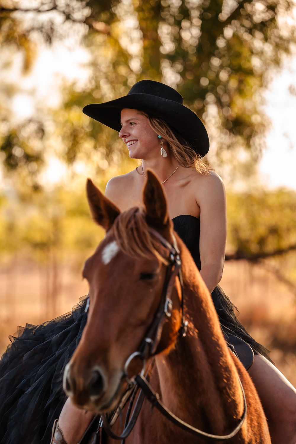 Woman in black hat riding a majestic brown horse during golden hour sunset creates a dreamy outdoor scene.