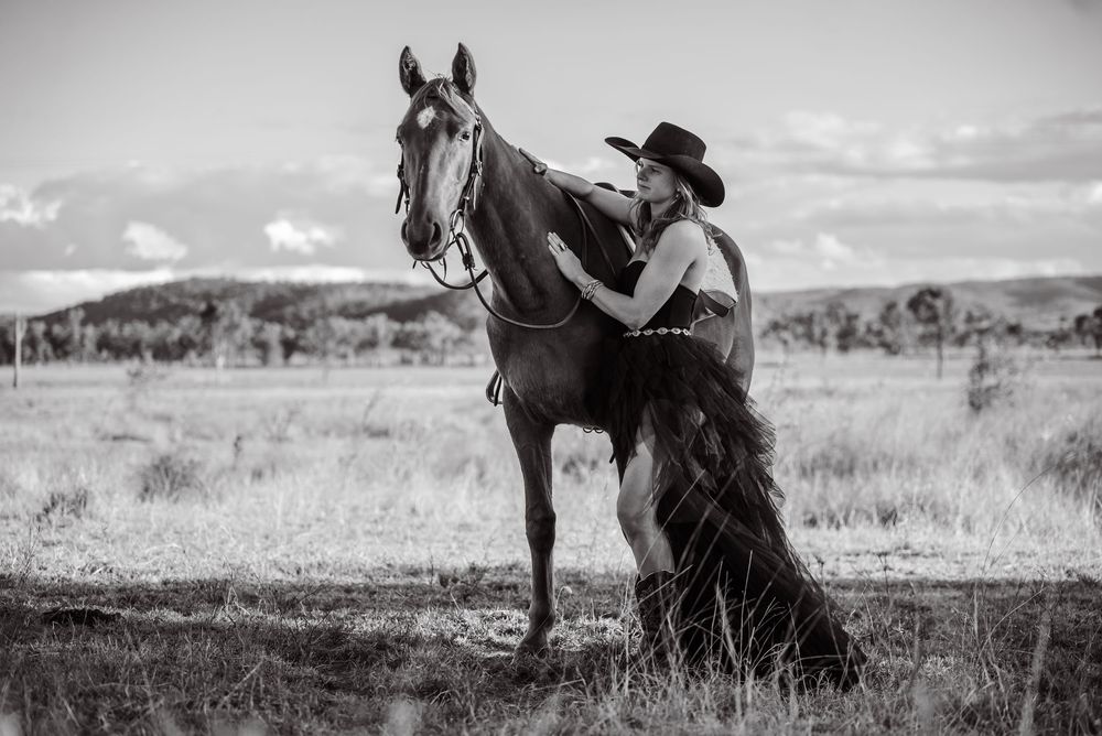 A black and white photo of a person in western attire standing with their horse in an open field.