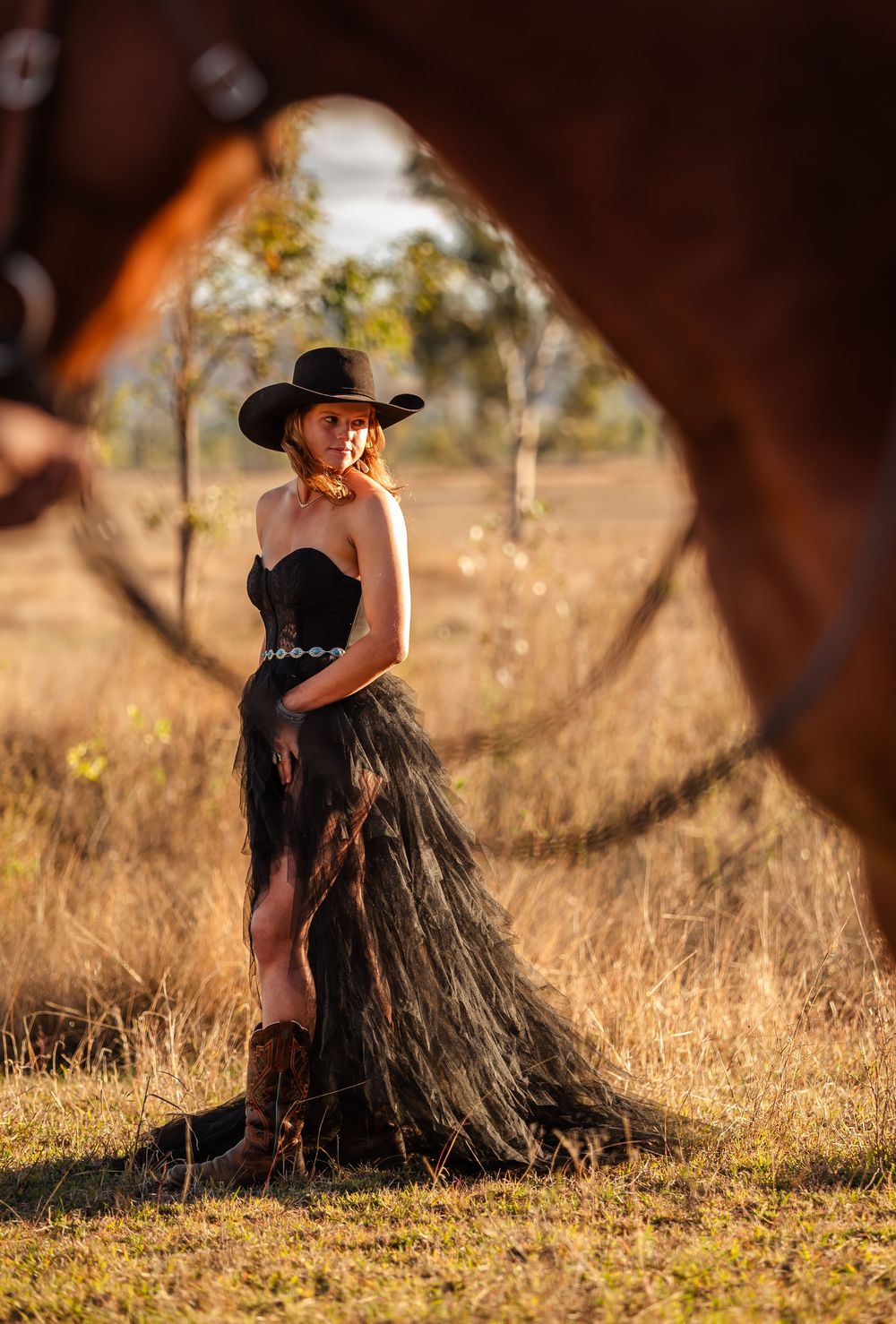 Elegant black tulle dress and cowboy hat silhouetted against golden field with rustic mirror frame at sunset.