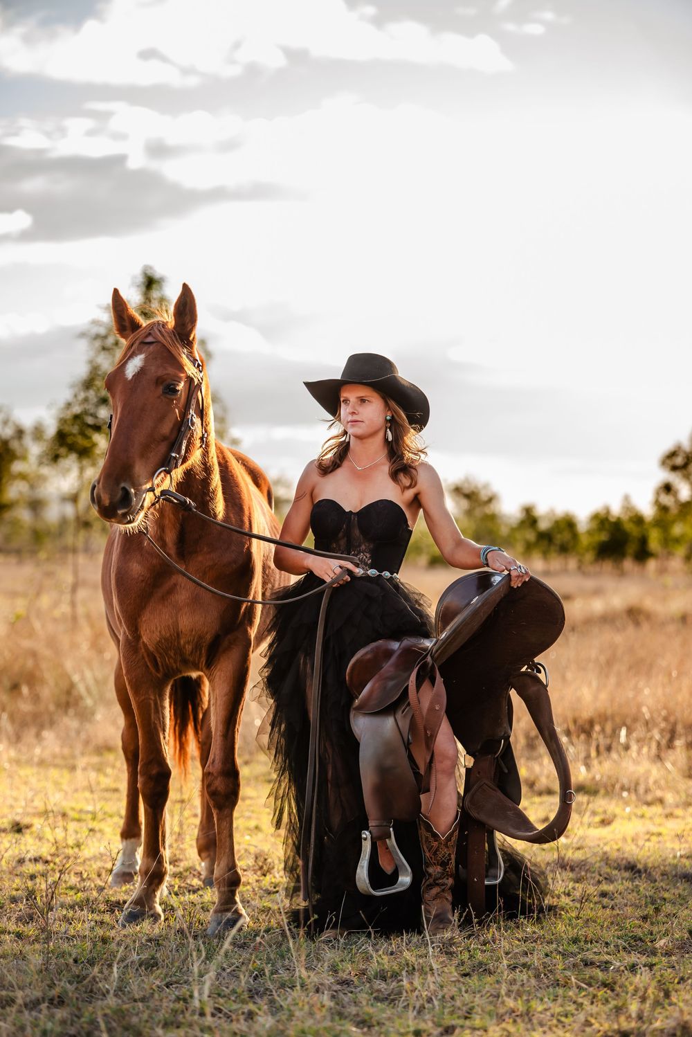 A person in black attire and cowboy hat sits beside a brown horse in a golden field at sunset.