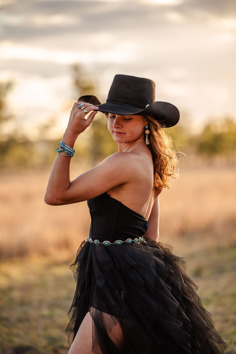 A figure in a black tulle dress and cowboy hat poses dramatically in golden sunlight at sunset.