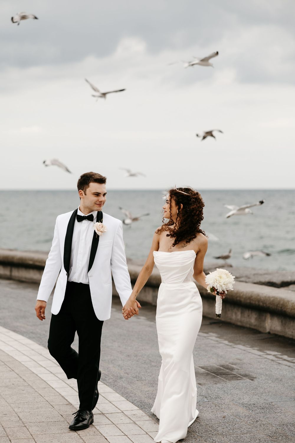 A couple walks along a waterfront pier while seagulls soar overhead in a cloudy coastal setting.