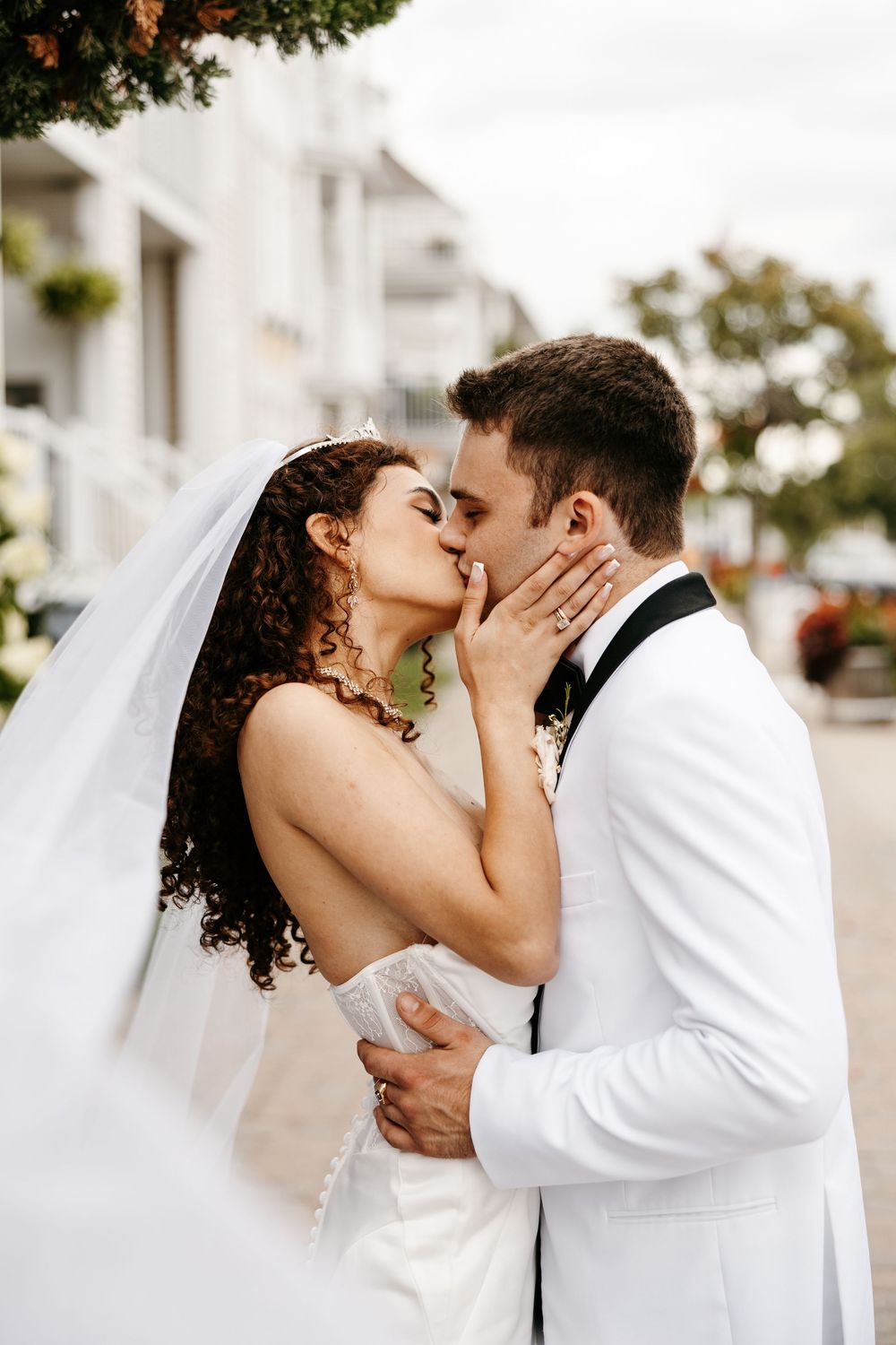 A romantic wedding moment captured outdoors with a bride in white gown and groom in white tuxedo jacket embracing.