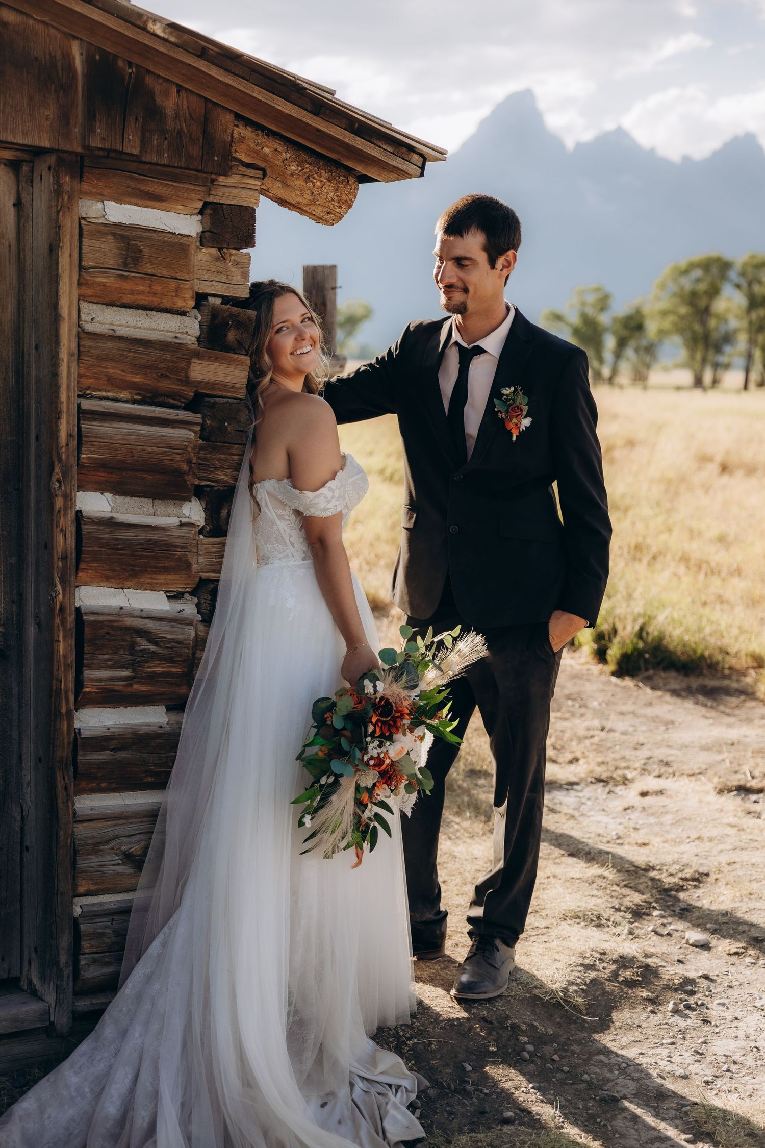 Couple shares romantic wedding day moments by rustic wooden cabin with majestic mountain backdrop in afternoon light.