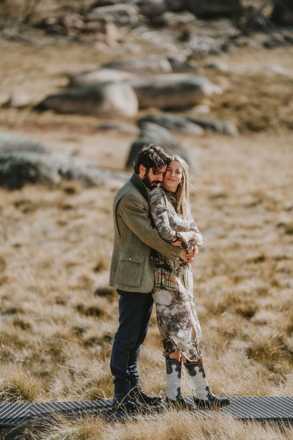 A couple shares a tender embrace while standing on a rustic wooden boardwalk in a mountainous outdoor setting.