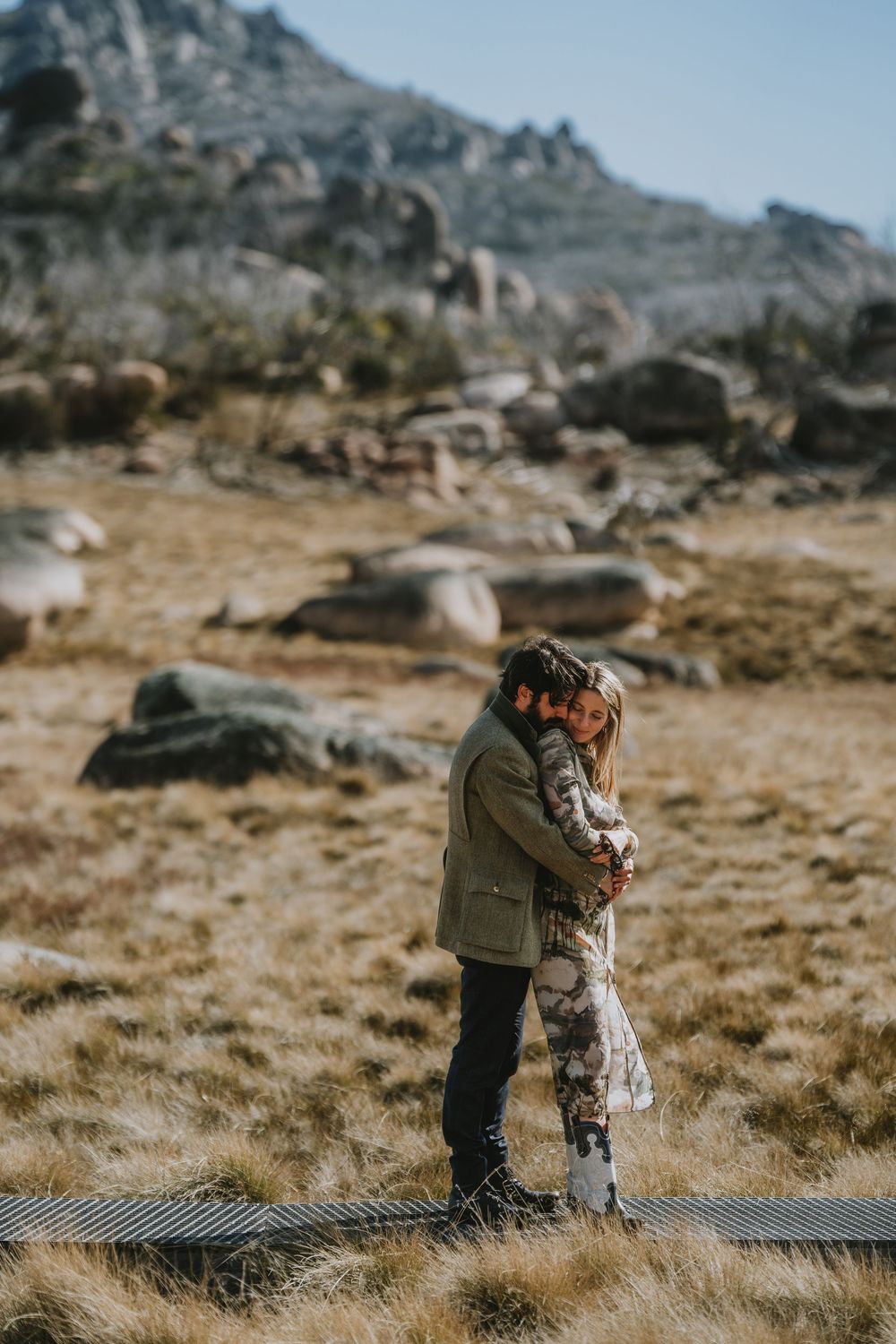Two people share an emotional moment on a wooden boardwalk surrounded by rocky mountain terrain and dry grass.