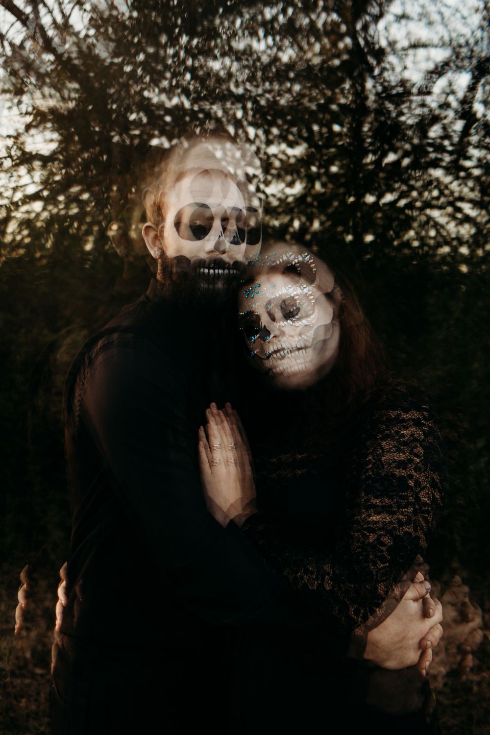 Ghostly double exposure portrait of couple in skull face paint embracing in dark wooded background.