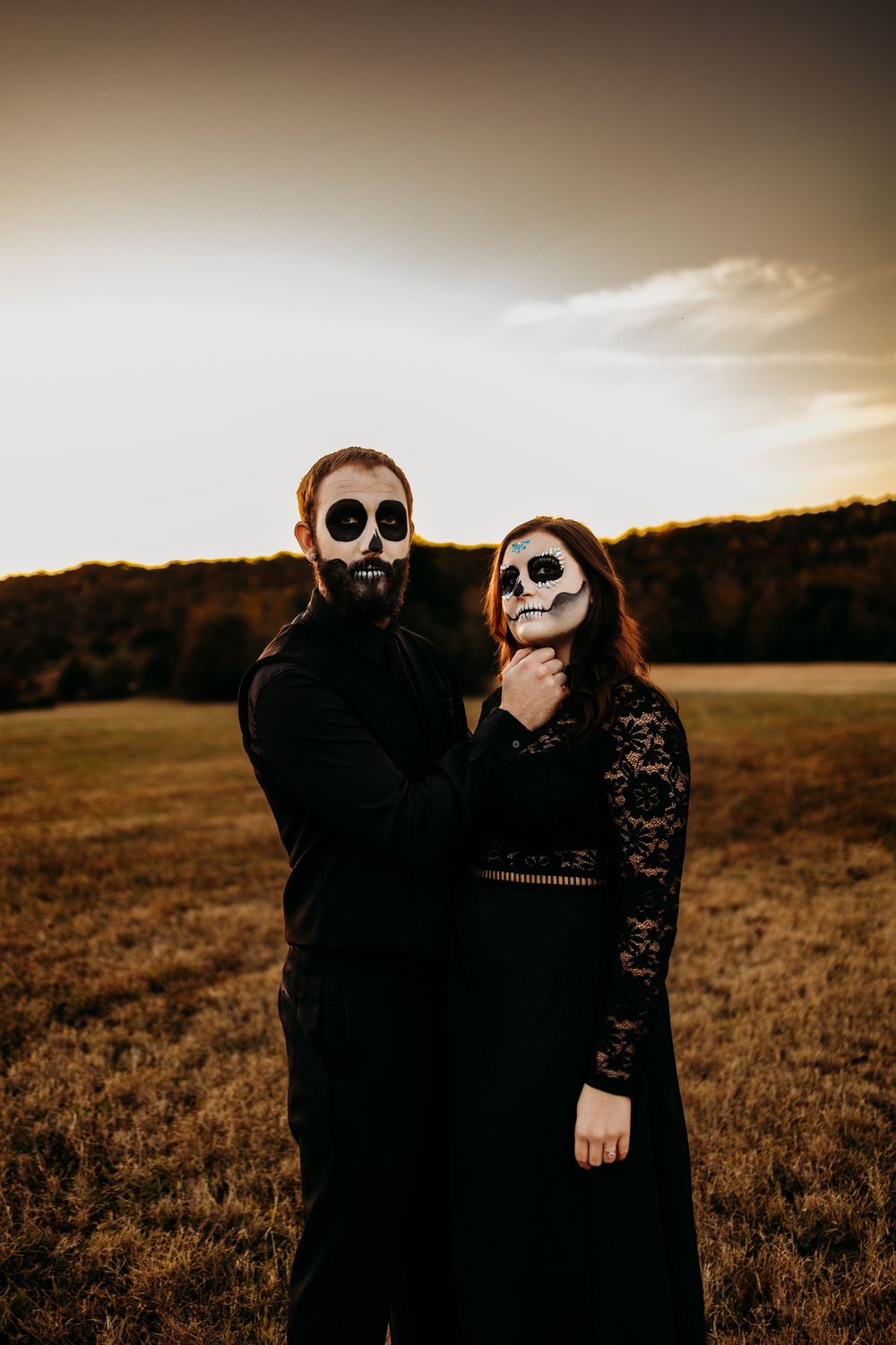 Couple in black attire with skull face paint stand in golden field at dusk under dramatic sky.