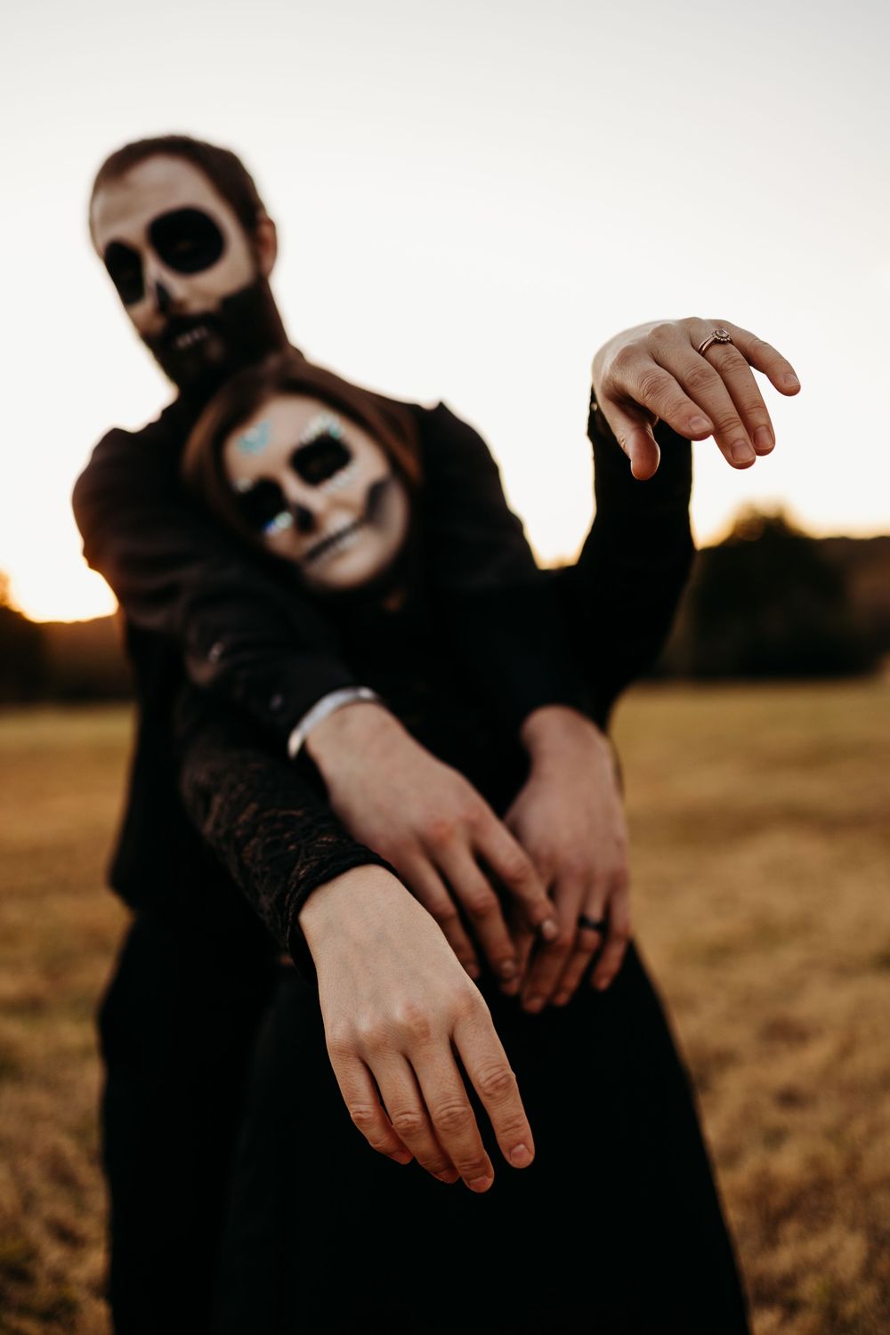 Couple in skeleton face paint and black clothing pose dramatically at sunset in an open field.