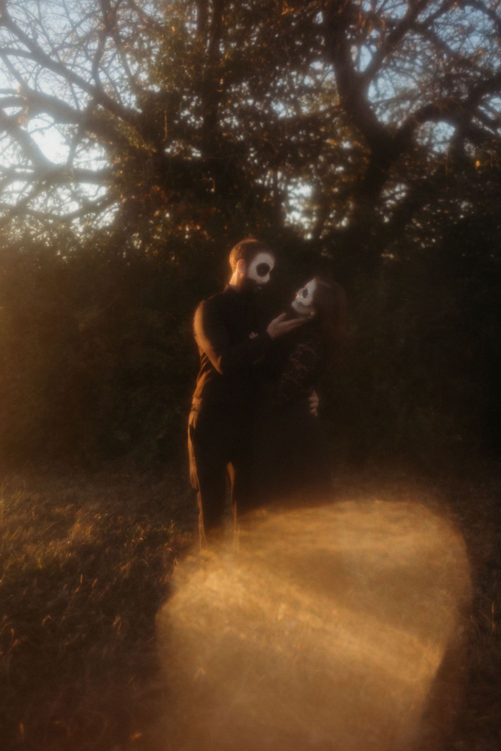 Couple in skull face paint embracing under bare tree in golden misty forest light.