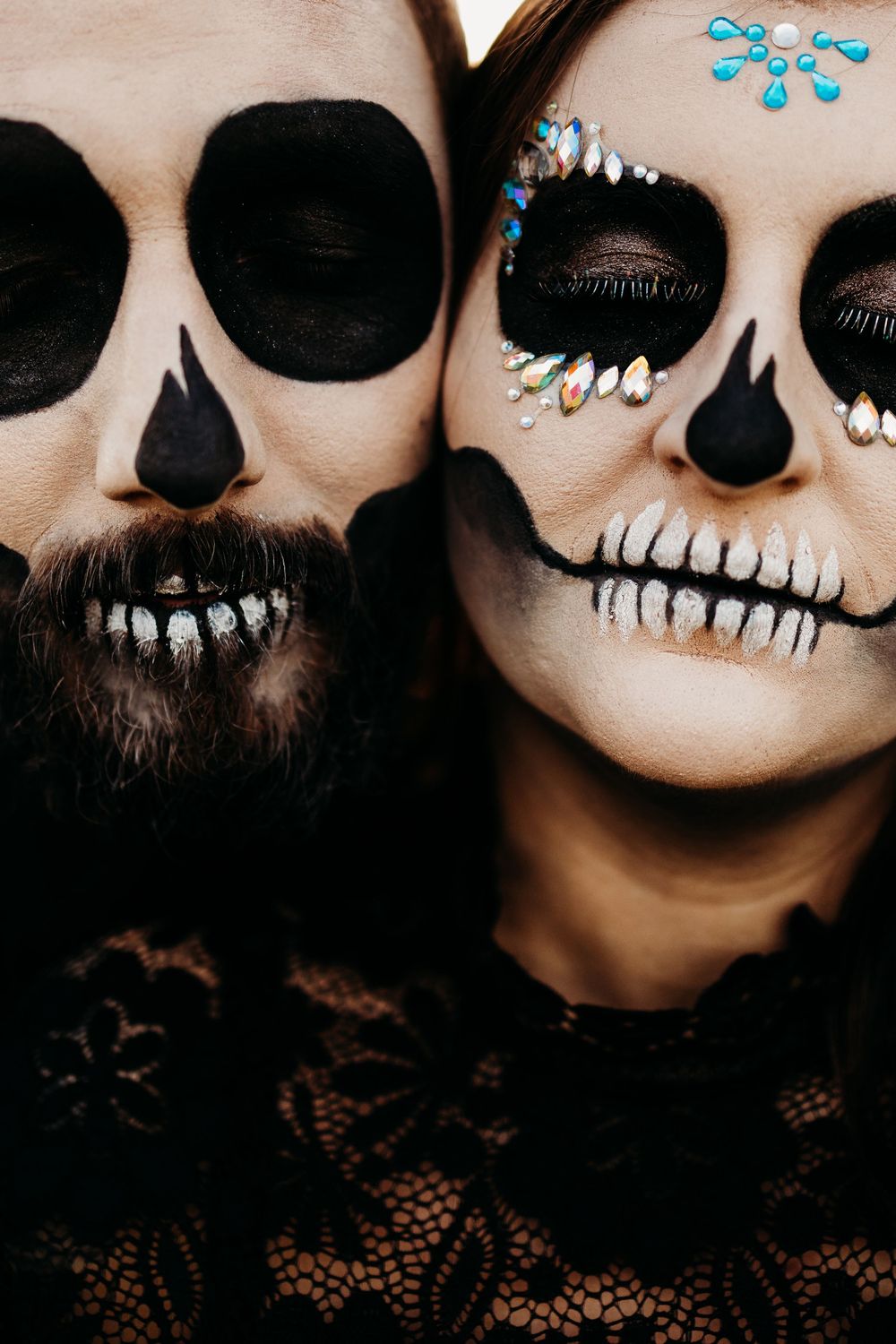 Close-up of couple wearing detailed skull face paint with jewel accents and black lace clothing.